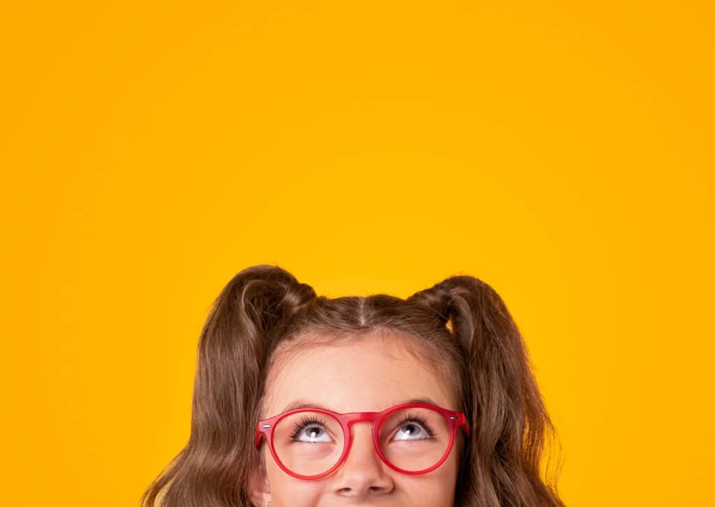 Positive teen girl with ponytails wearing stylish red framed eyeglasses looking up on yellow background.
