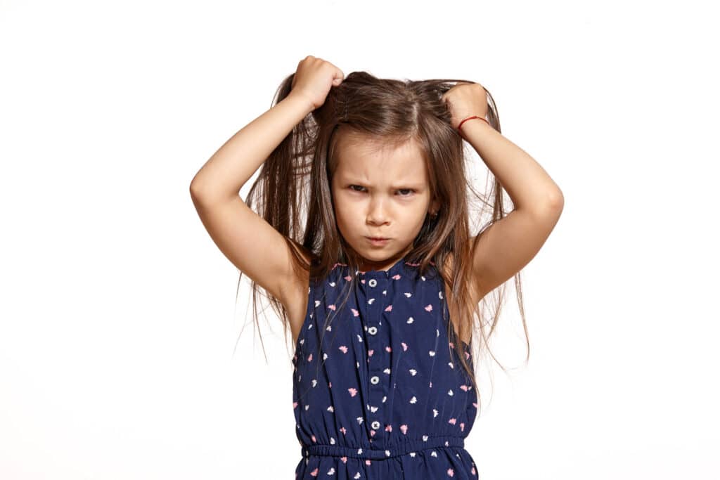 A young girl with long brown hair, wearing a blue polka-dot dress, pulling her hair and looking frustrated with a serious expression.